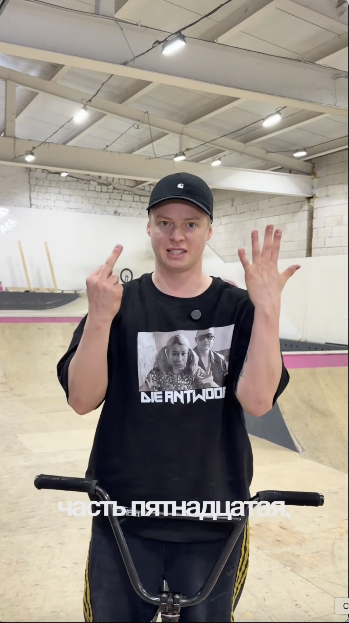 Kid-led camp invitation opener: a young-looking founder in a black cap and Die Antwoord t-shirt standing behind BMX handlebars in an indoor skatepark, hands raised mid-talk, caption "часть пятнадцатая" on screen.