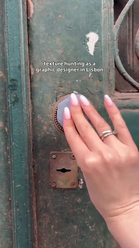 A manicured hand presses a disc of grey clay against a weathered green ornate door with a decorative metal knob, captioned "texture hunting as a graphic designer in Lisbon".