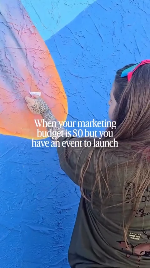 A woman seen from behind hand-paints a large mural, bold orange over a deep blue wall, captioned "When your marketing budget is $0 but you have an event to launch".