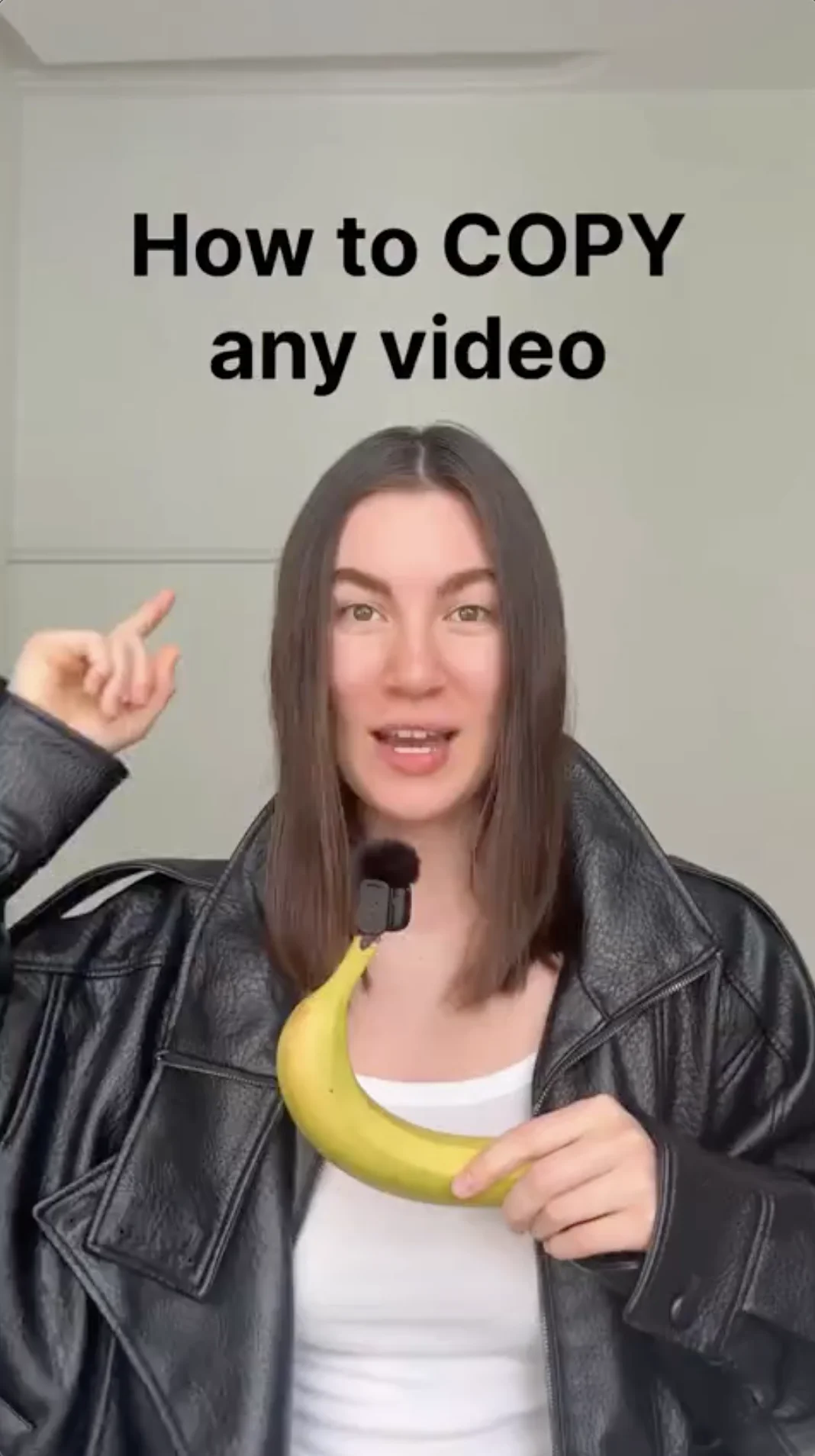 Vertical opening frame from alinamerkelcoach's tutorial. The bold black headline "How to COPY any video" sits top-center over a plain wall; the creator, in a leather jacket, holds a banana with a lavalier microphone clipped to it.