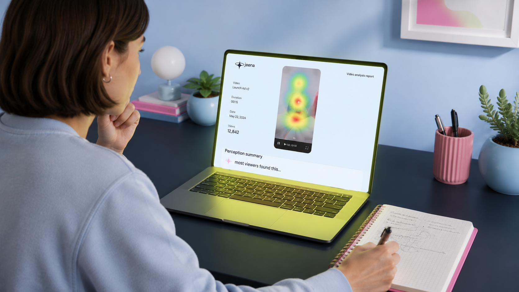 A marketer at a laptop in a softly lit home office, viewed from over her shoulder, reviewing a Jeena video analysis report. The laptop screen shows the jeena wordmark in the upper-left, a vertical video frame with a hot red-green attention heatmap overlay in the centre, and a "Perception summary: most viewers found this..." line beneath. Video metadata on the left reads "Video: Launch Ad v2 · Duration 00:15 · Views 12,842". A spiral notebook with a pink edge sits on the desk to her right, a pen mid-note in her hand. Pencil cup and plant flank the laptop.