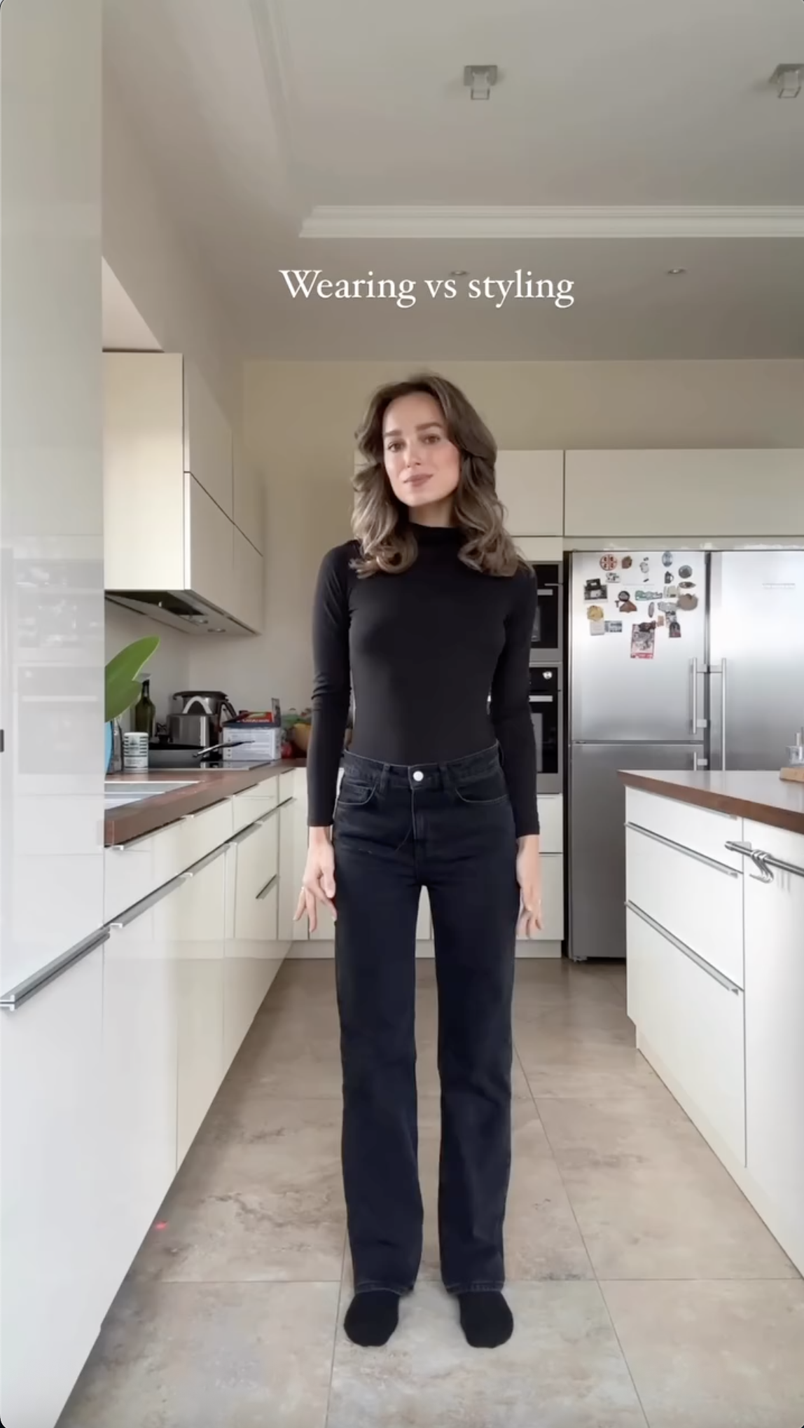 Vertical screenshot from the 112k-view "wearing vs styling" video. A woman in a black turtleneck and high-waisted black jeans in a kitchen, with the "Wearing vs styling" title set in larger serif at the top.